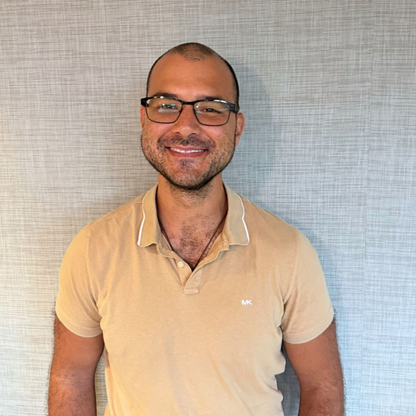 Smiling man with short hair and glasses wearing a beige polo shirt, standing against a light gray textured wall.