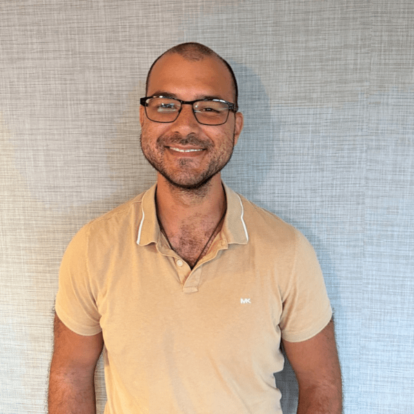Smiling man with short hair and glasses wearing a beige polo shirt, standing against a light gray textured wall.