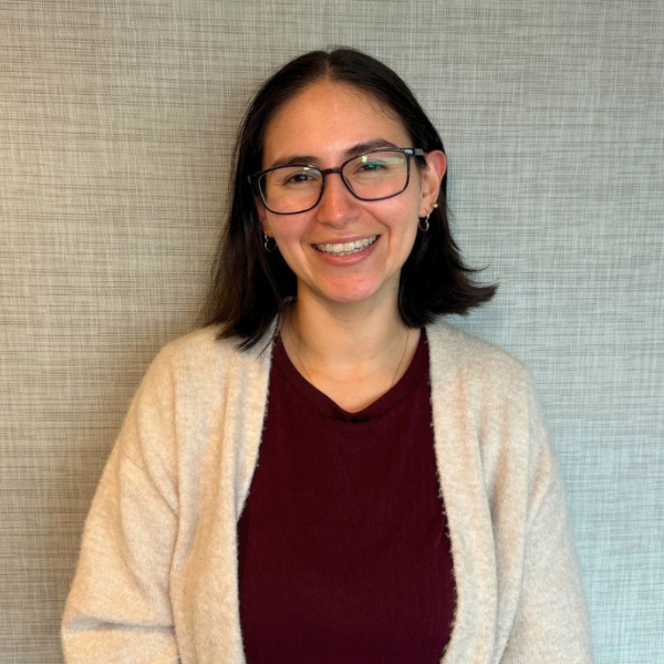 Smiling woman with glasses and shoulder-length dark hair, wearing a burgundy top and beige cardigan, standing against a textured gray wall.