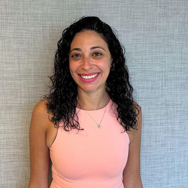Smiling woman with shoulder-length curly black hair wearing a light pink sleeveless top, posed against a gray textured wall.