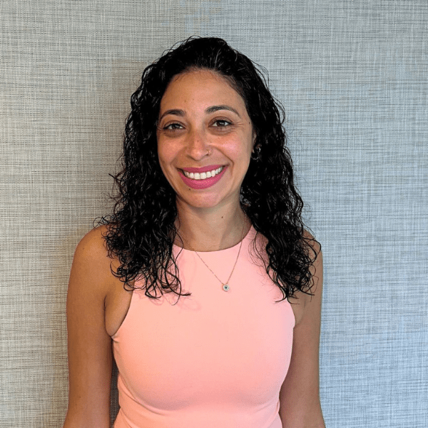 Smiling woman with shoulder-length curly black hair wearing a light pink sleeveless top, posed against a gray textured wall.