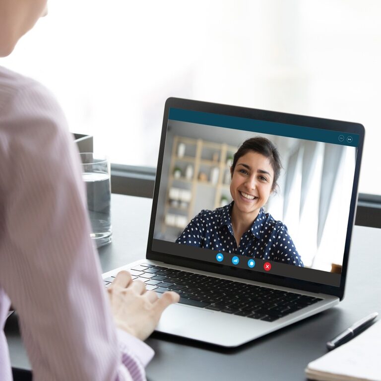 Indian girl communicate with friend on-line by video call, pc screen view over female shoulder. Mental health expert online therapy, colleagues work on common project use videoconferencing app concept